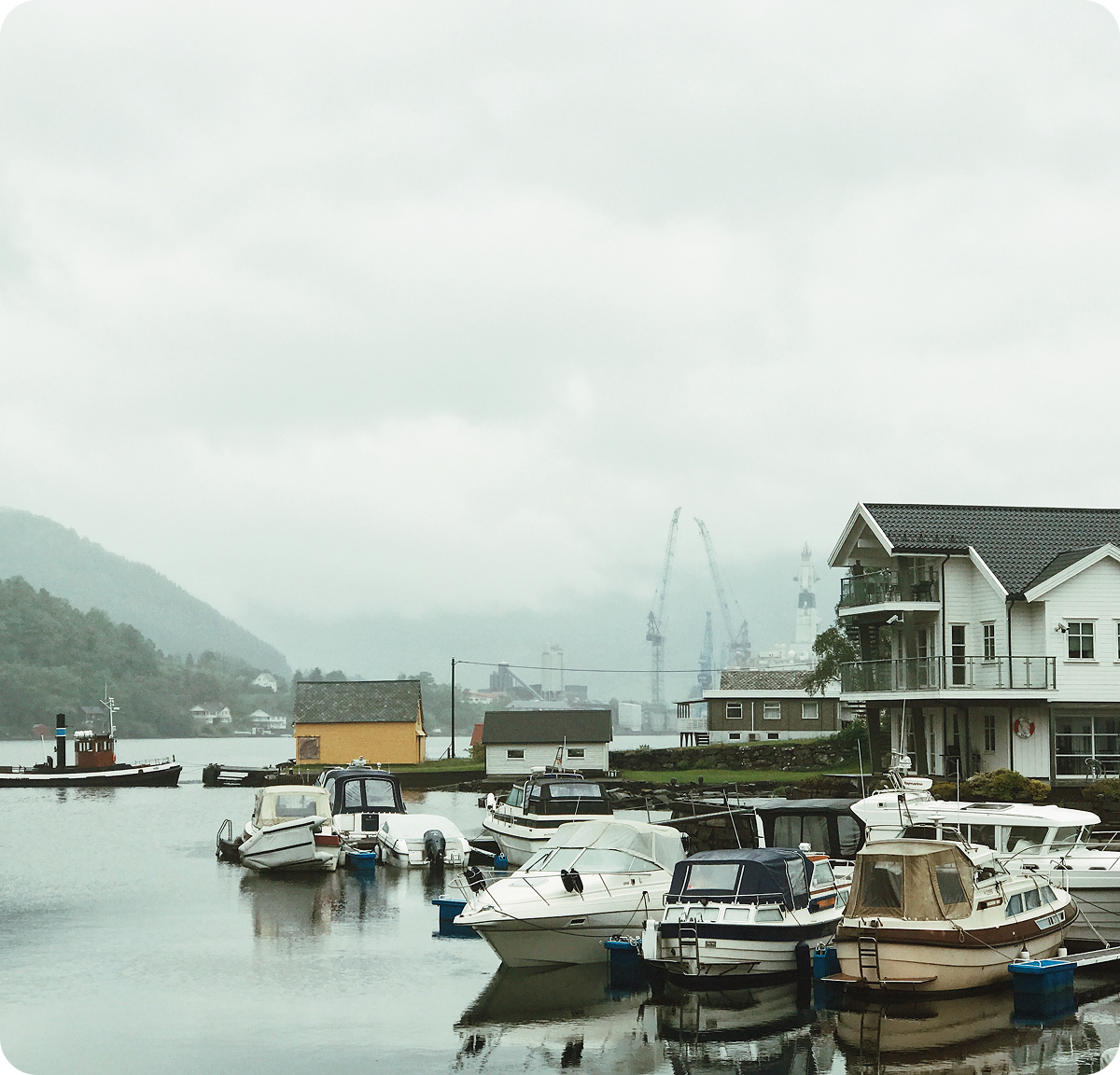 Boats docked at a calm harbor under cloudy skies with misty hills in the background.